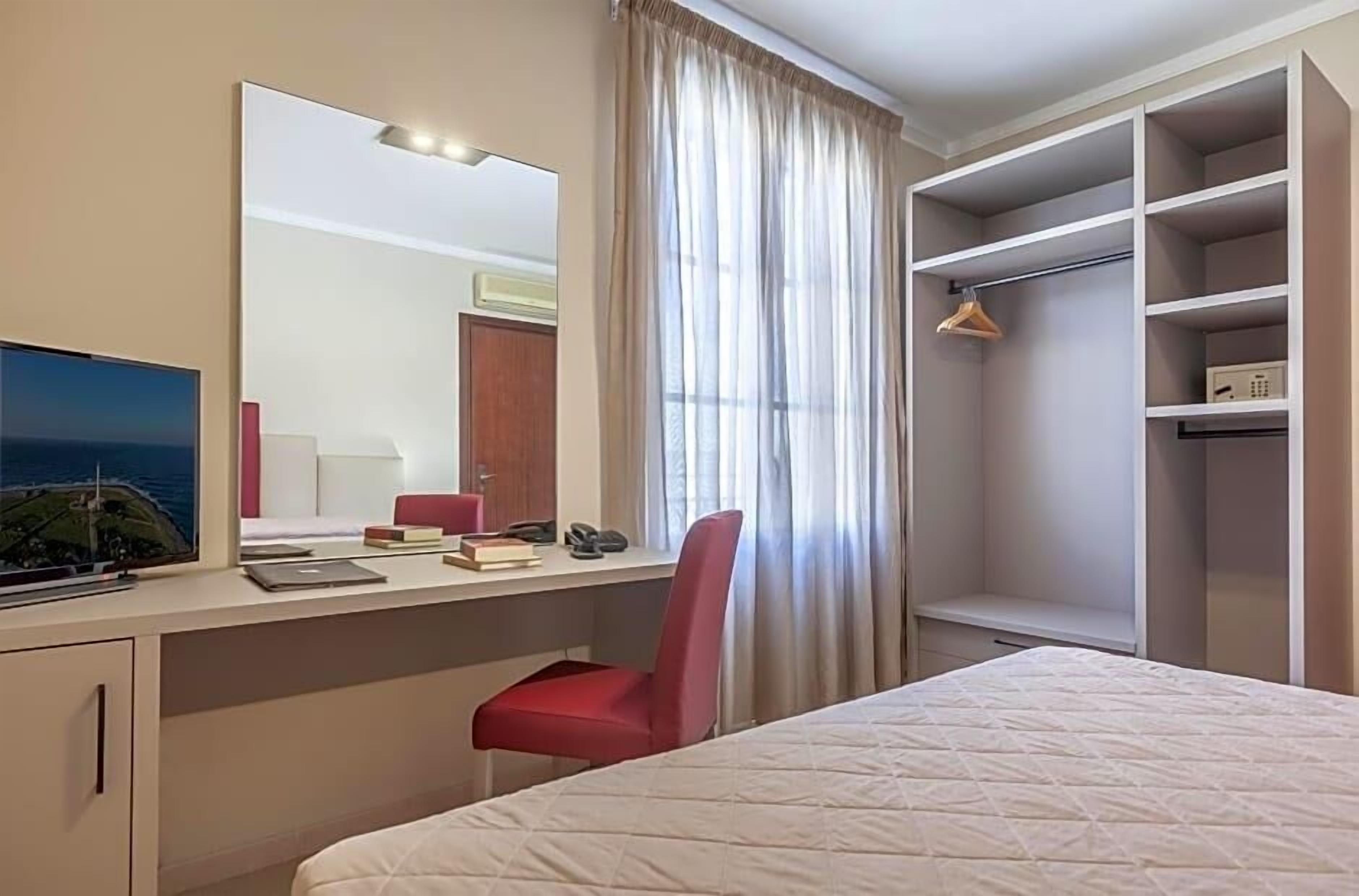 Habitación de hotel con cama, escritorio con computadora, silla, espejo y armario.