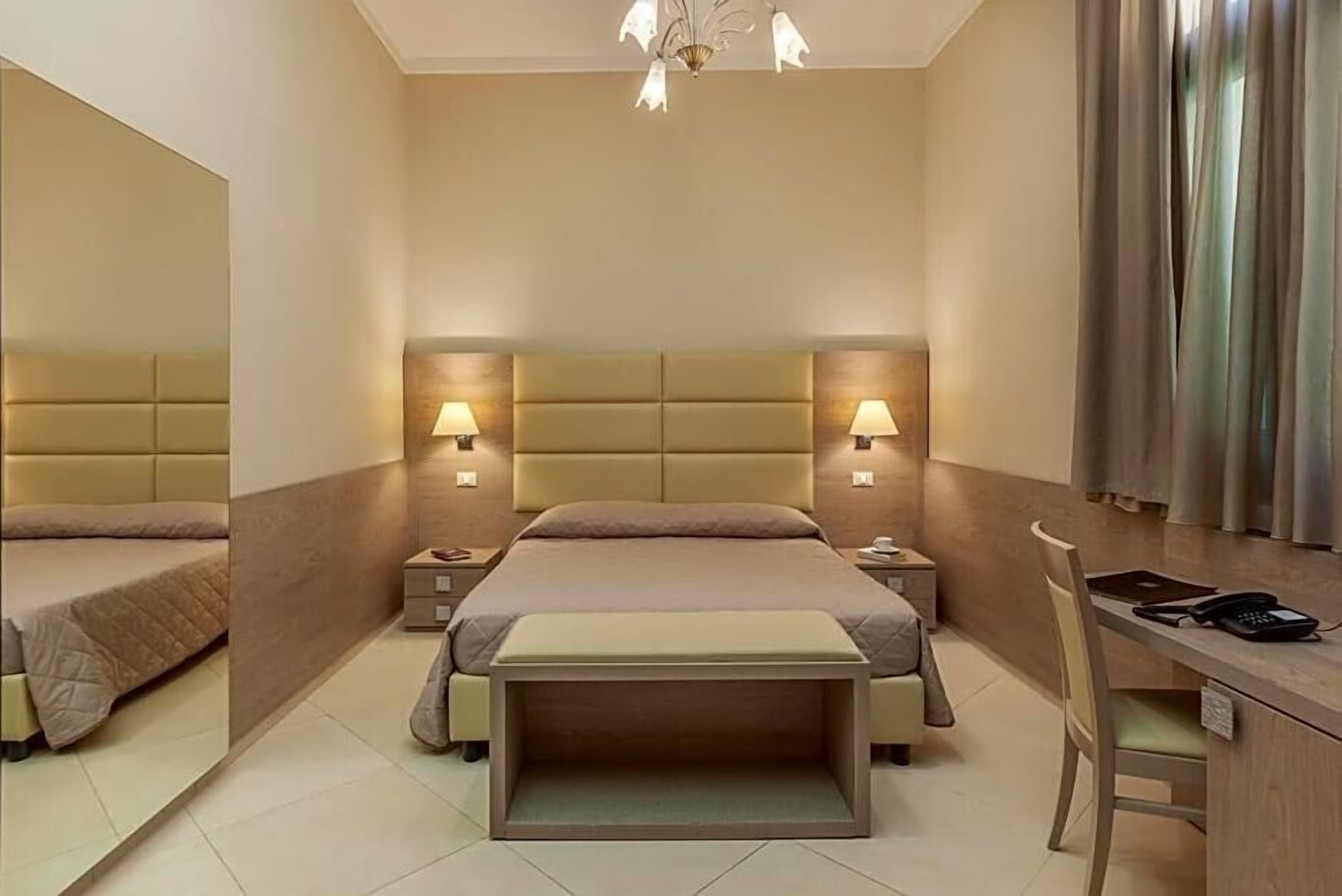 Quarto de hotel com uma cama grande, mesas de cabeceira com luminárias, uma escrivaninha com cadeira e um espelho.