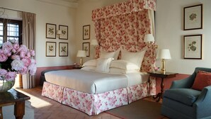 Een slaapkamer met een groot bed, bloemen gordijnen, ingelijste botanische prints aan de muur en een bloemenvaas.