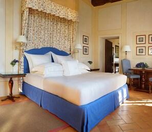 A large bed with a blue headboard and white bedding, flanked by two bedside tables with lamps, in a room with a floral curtain and framed pictures on the wall. - Castello Banfi (Montalcino)