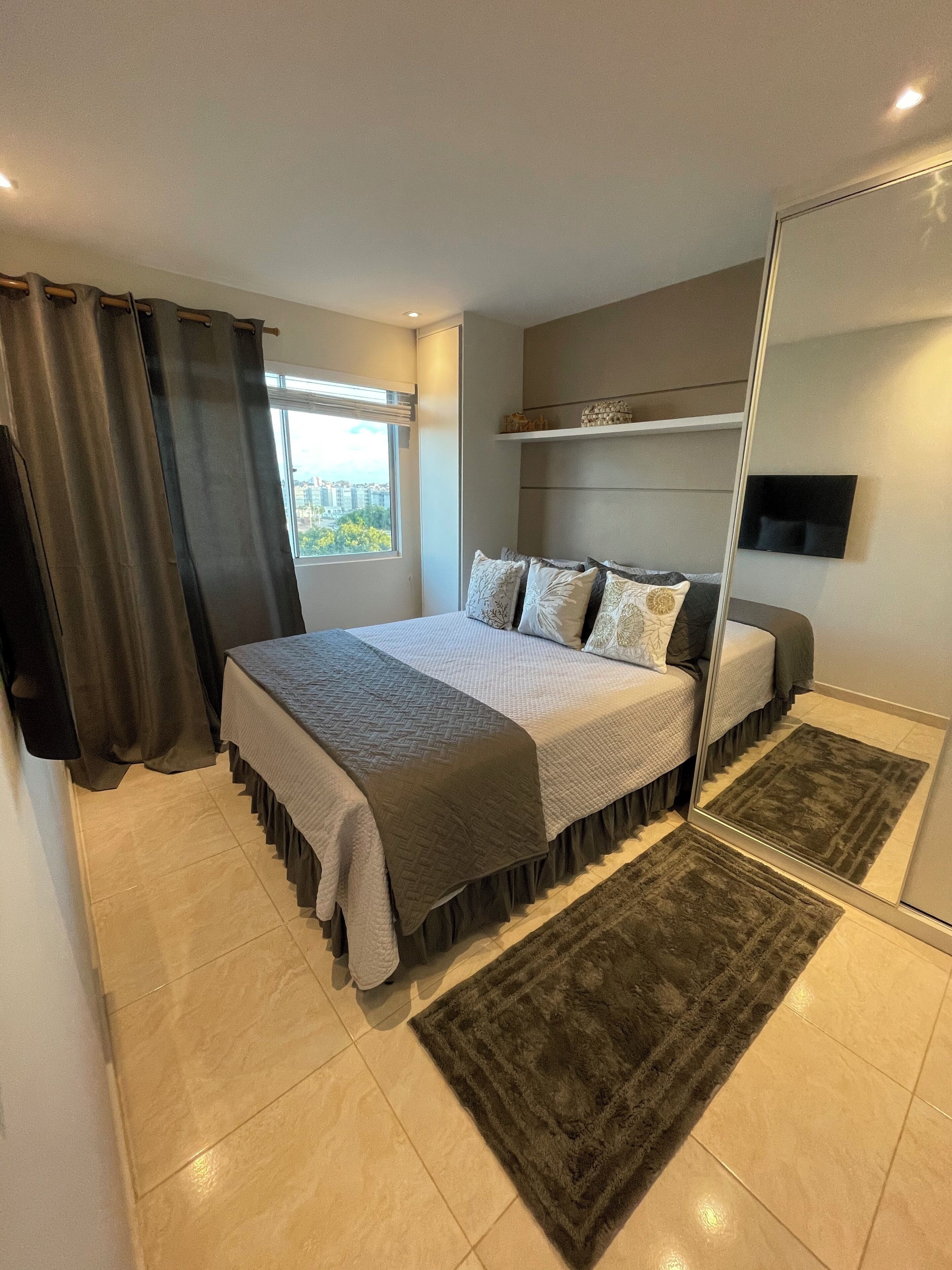 A modern bedroom with a large bed, a flat-screen TV mounted on the wall, and a view of the city through the window.