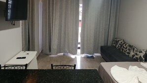 A hotel room with a bed, a sofa, a TV mounted on the wall, a small table with a remote control, and a window with curtains. - Studio apartment with bedroom, kitchen, and bathroom, comfortable and cozy, independent. (Contagem)