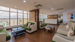 Un moderno vestíbulo de hotel con ventanas amplias, sofás verde y blanco, una mesa de centro y un mostrador de recepción con un reloj y cuadros enmarcados.