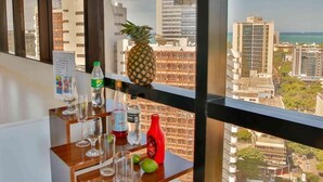Una mesa con una piña, botellas y vasos, con vista a un paisaje urbano con edificios y al mar.