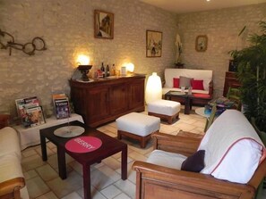 Uma sala de estar acolhedora com parede de pedra, um armário de madeira, um sofá branco, uma poltrona de madeira e uma mesa de centro com um jogo americano 'BERRY'.