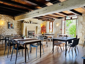 Een rustiek interieur met zichtbare houten balken, stenen muren en een open haard. Tafels en stoelen staan klaar voor het diner.