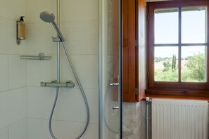 En brusekabine med brusehoved, sæbedispenser og vindue med udsigt til grøn natur.