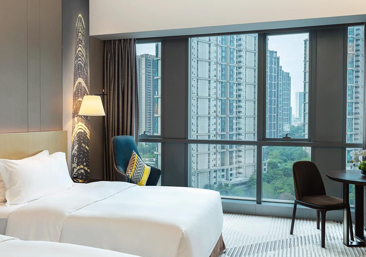 Une chambre d’hôtel avec un grand lit, une chaise, une petite table et une vue sur de hauts immeubles, visible à travers de grandes fenêtres.