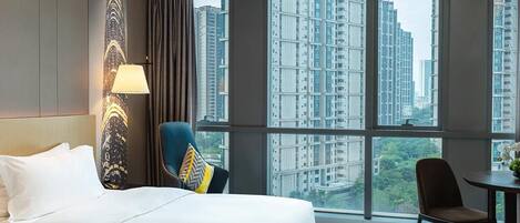 Une chambre d’hôtel avec un grand lit, une chaise, une petite table et une vue sur de hauts immeubles, visible à travers de grandes fenêtres.