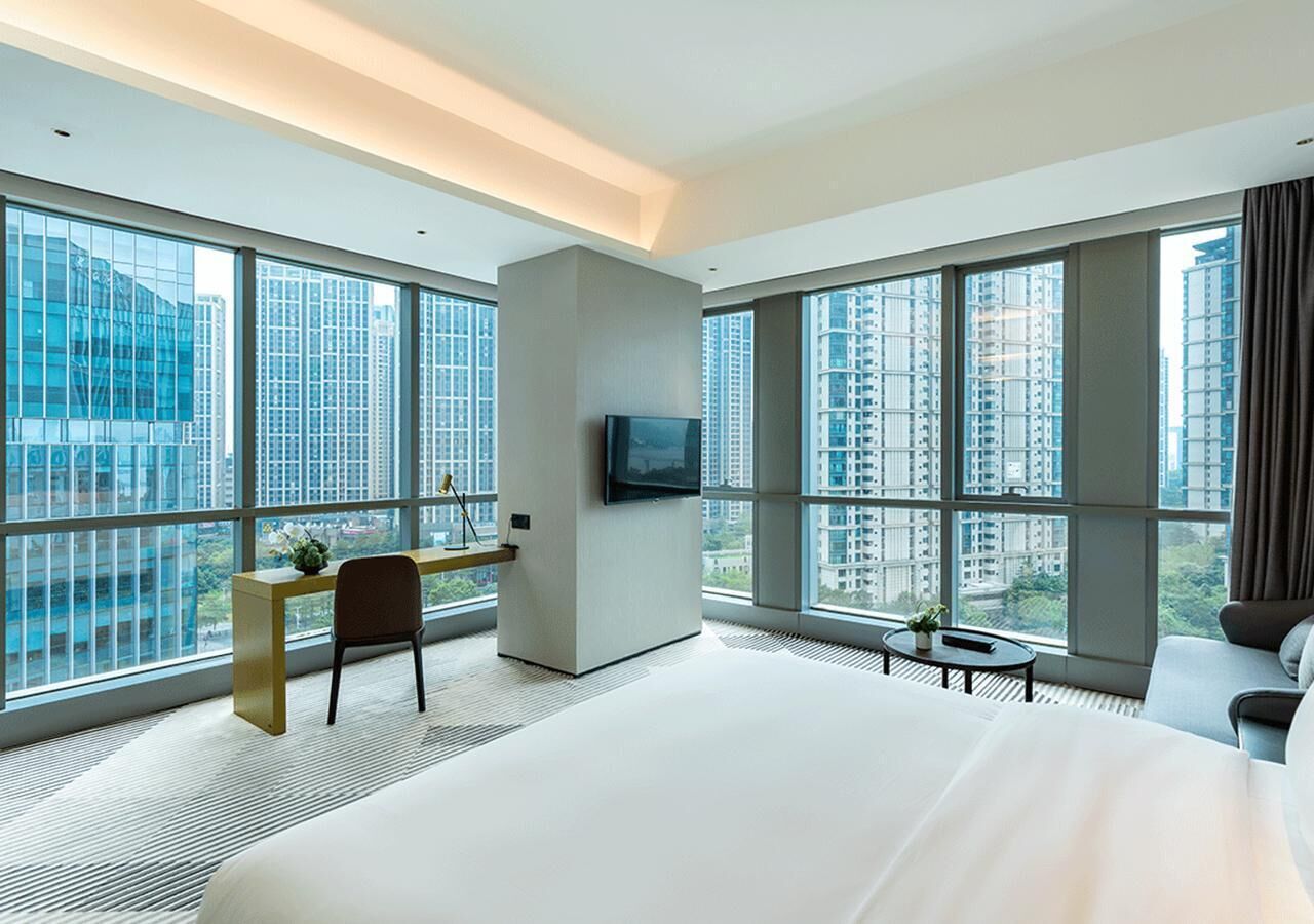 Ein modernes Hotelzimmer mit großen Fenstern, einem Flachbildfernseher, einem Bett, einem Schreibtisch und einem Sessel.
