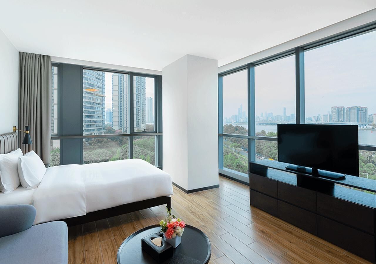 A modern hotel room with a large bed, a sofa, a TV, and a city view through large windows.