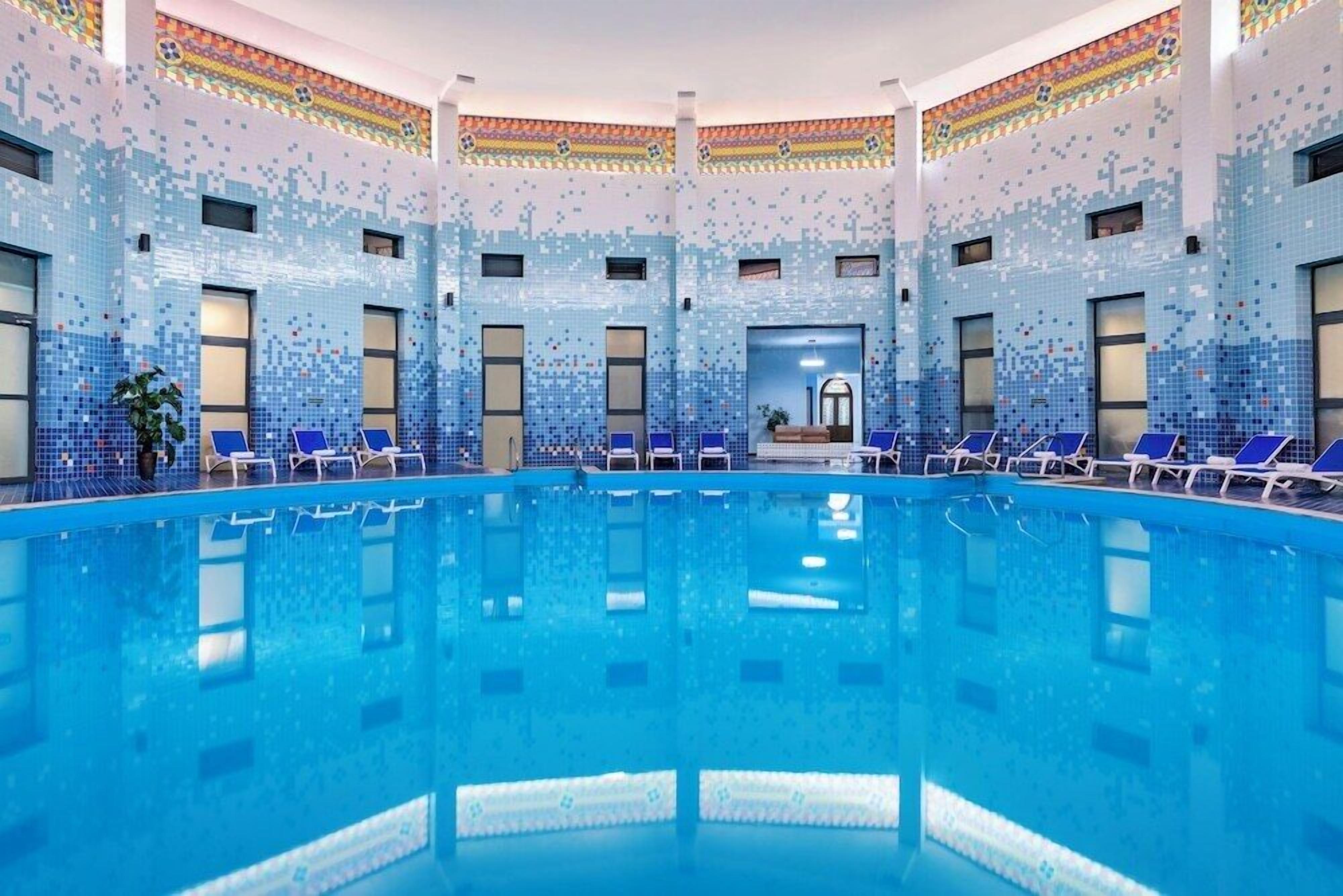 A large indoor swimming pool with blue-tiled walls and multiple lounge chairs around the perimeter.