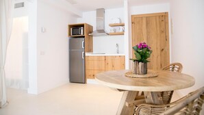 A compact kitchen with a microwave, stove, and wooden cabinets. A dining table with chairs is in the foreground. - Sa Cala Suites (Formentera)