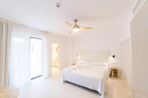A hotel room with a bed, a ceiling fan, a bedside table, and a view of the outside. - Sa Cala Suites (Formentera)