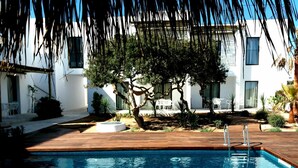 A swimming pool with a wooden deck, surrounded by white buildings and trees. - Sa Cala Suites (Formentera)