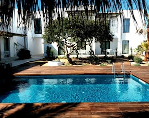 A swimming pool with a wooden deck, surrounded by white buildings and trees. - Sa Cala Suites (Formentera)