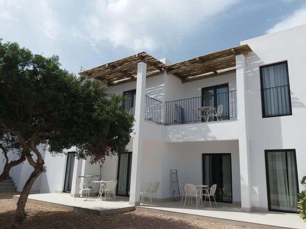 A modern two-story building with a covered patio area, outdoor seating, and a tree. - Sa Cala Suites (Formentera)
