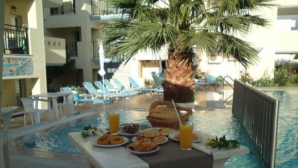 Une table placée au bord de la piscine, avec de la nourriture et des boissons, entourée de chaises longues et d’un palmier.