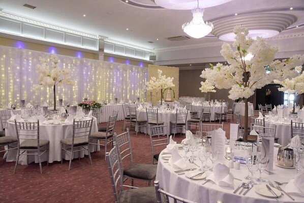 Uma sala de banquetes com mesas redondas preparadas para refeições, centros de mesa florais brancos e um candelabro.