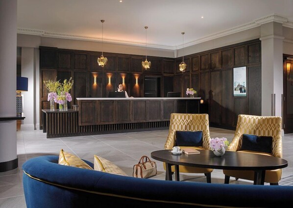 A modern hotel lobby with a reception desk, dark wood paneling, a blue sofa, and two patterned armchairs. - Carrigaline Court Hotel AND Leisure Centre (Carrigaline)