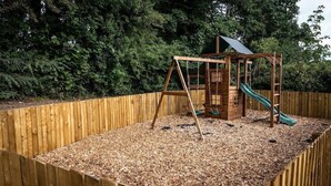 A wooden play structure with a slide and swings, surrounded by a wooden fence and a wooded background. - The Vicarage (Crewe)