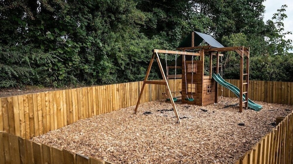 Una estructura de juego de madera con un tobogán y columpios, rodeada por una valla de madera y un fondo de bosque.