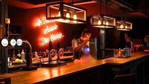 A bar area with a neon sign reading "The Bishop," a bartender pouring a drink, and a selection of bottles and glasses on the counter. - The Vicarage (Crewe)