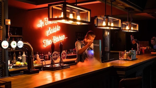 A bar area with a neon sign reading "The Bishop," a bartender pouring a drink, and a selection of bottles and glasses on the counter. - The Vicarage (Crewe)