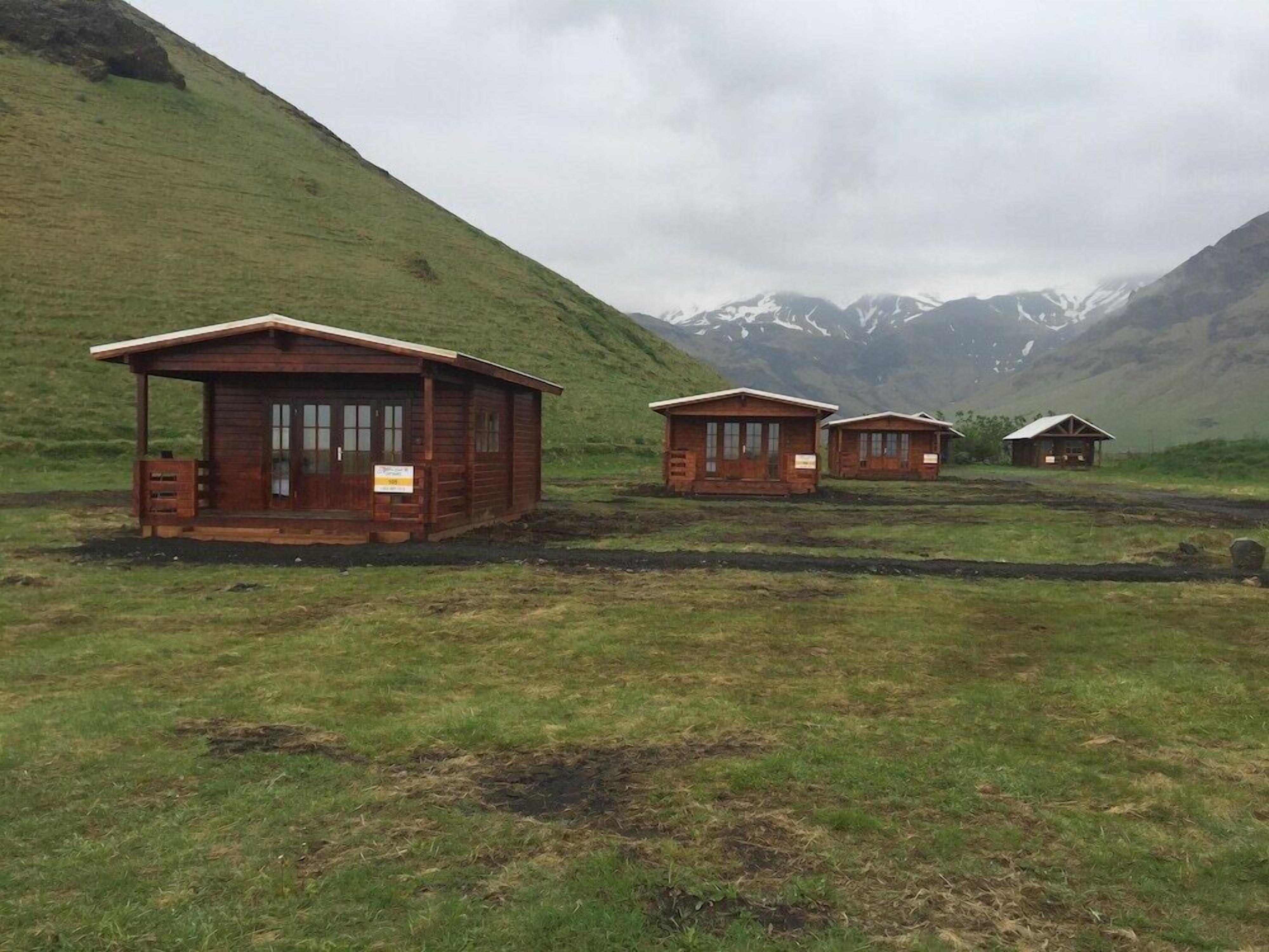 Un hilera de cabañas de madera en un campo cubierto de hierba, con montañas al fondo.