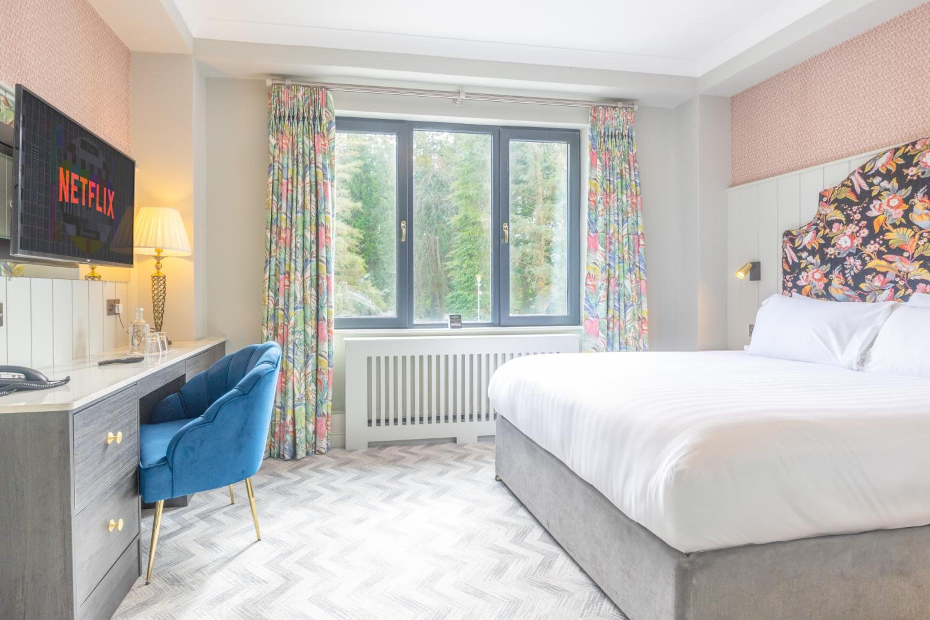 A hotel room with a bed, a desk with a computer, a chair, a window with curtains, and a wall-mounted TV.