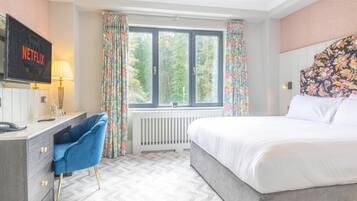 A hotel room with a bed, a desk with a computer, a chair, a window with curtains, and a wall-mounted TV.
