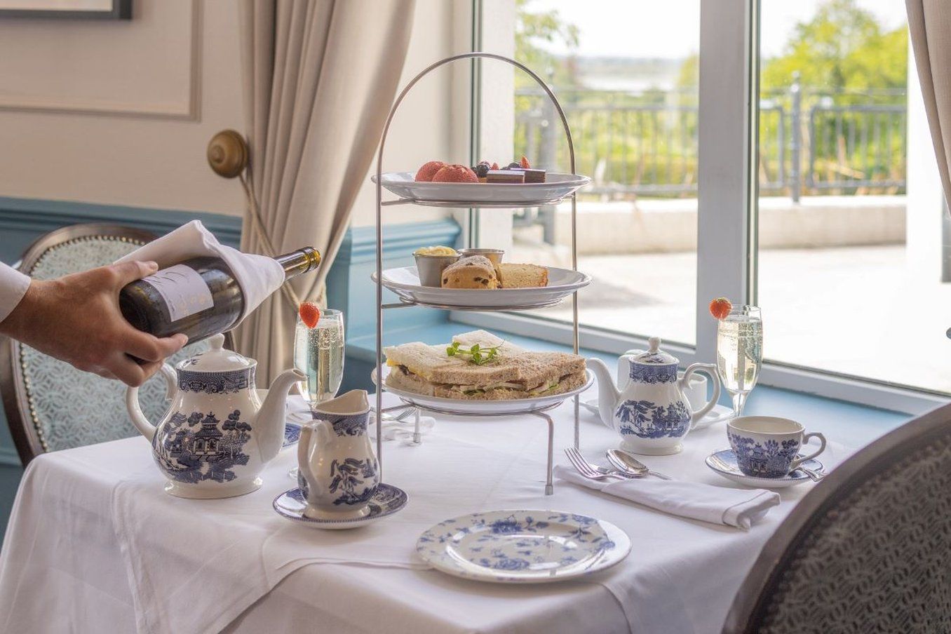 A table set for a tea service with a tiered tray of sandwiches, a teapot, cups, and a bottle being poured.