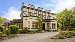 A traditional Tudor-style building with a symmetrical facade, a central entrance with an arch, and a well-maintained garden with various shrubs and a fountain. - Brook Hall Hotel (Ellesmere Port)