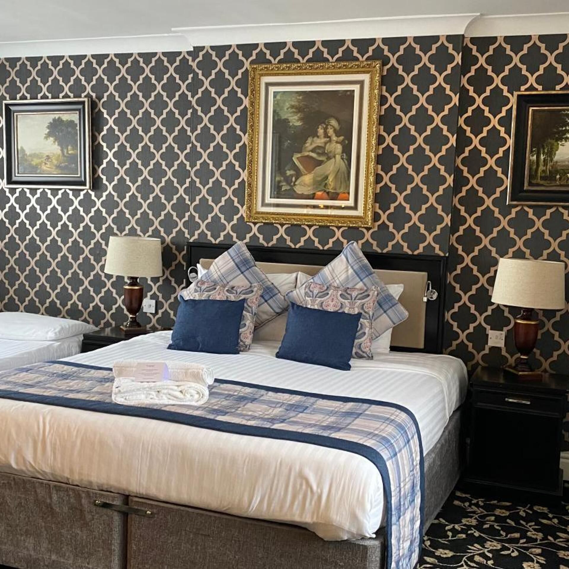 A hotel room with a large bed, two bedside tables with lamps, a patterned wallpaper, and framed artwork on the walls.