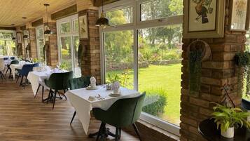 Un restaurante con mesas dispuestas para comer, ventanas amplias con vistas a un jardín y plantas colgantes.