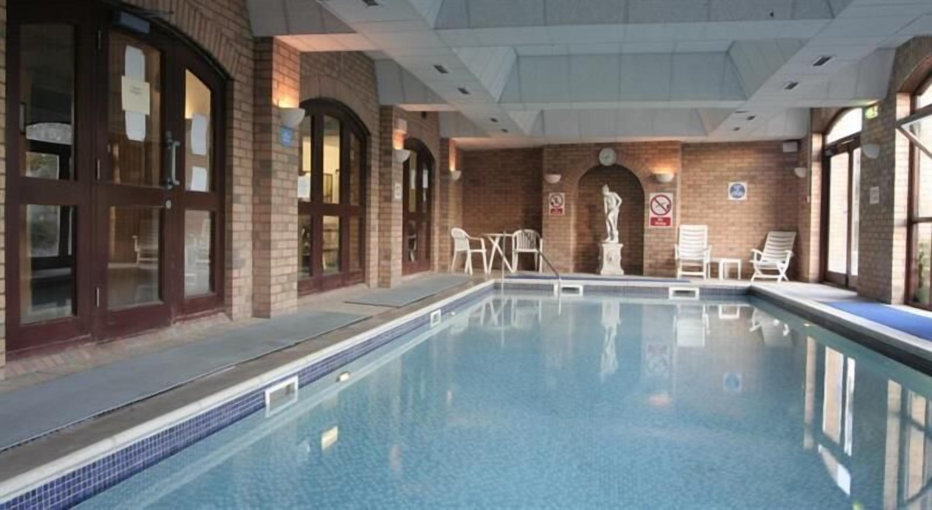 Una piscina interior con agua cristalina, rodeada de muros de ladrillo y ventanas amplias. Hay sillas de descanso y una estatua cerca de la piscina.