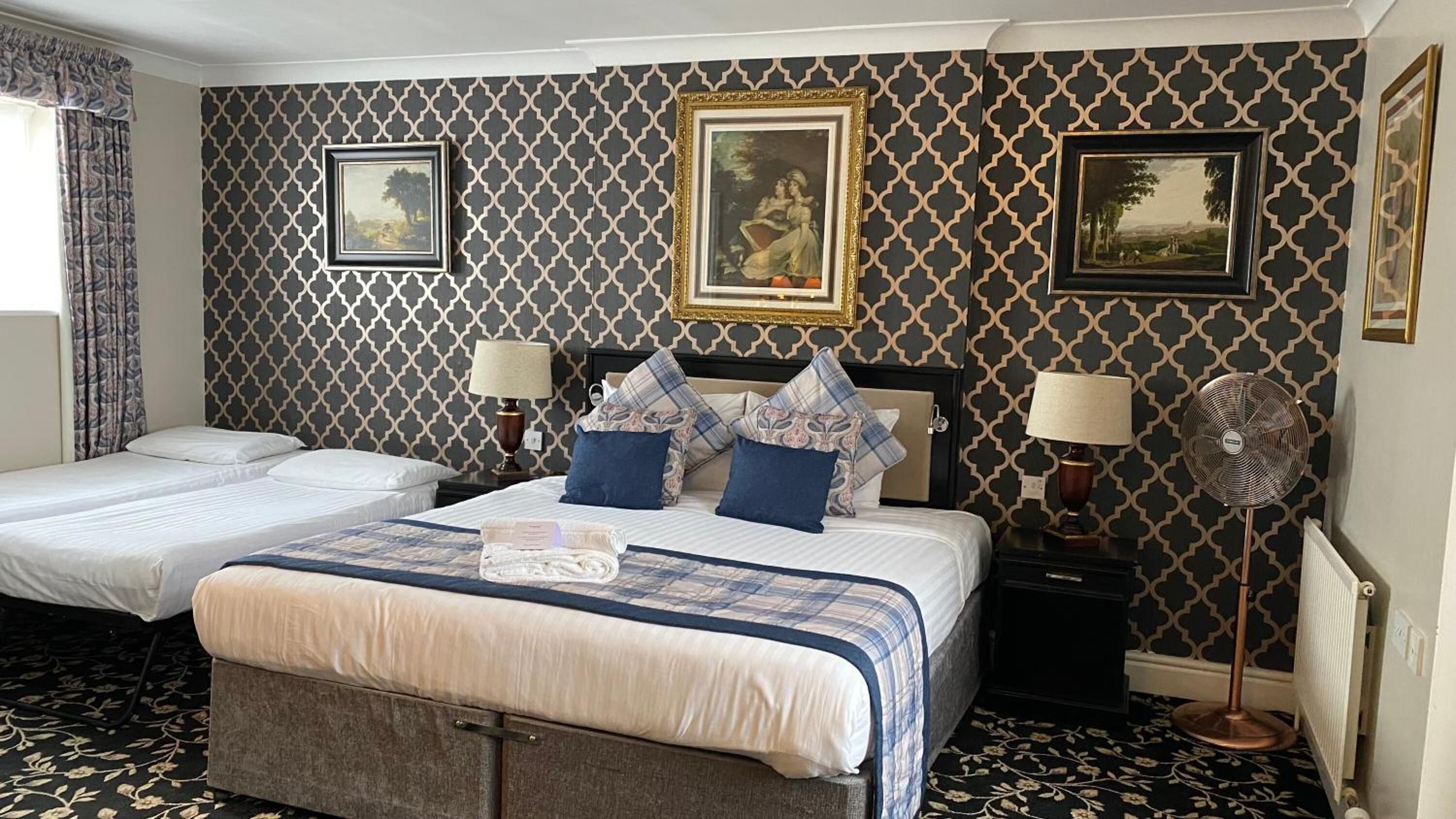 A hotel room with two beds, a large bed with a blue and white checkered bedspread, and two smaller beds. There are framed pictures on the wall, a standing fan, and a window with curtains.