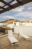 Terraza en la azotea con dos sillones blancos, una pequeña mesa redonda y una botella de vino. La terraza ofrece vistas a un paisaje montañoso y edificios blancos.