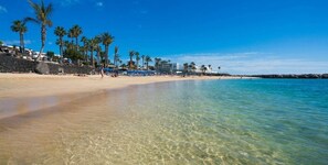 A beach with clear shallow water, palm trees, and a stone wall. - Playa Blanca Beach Mate (Yaiza)