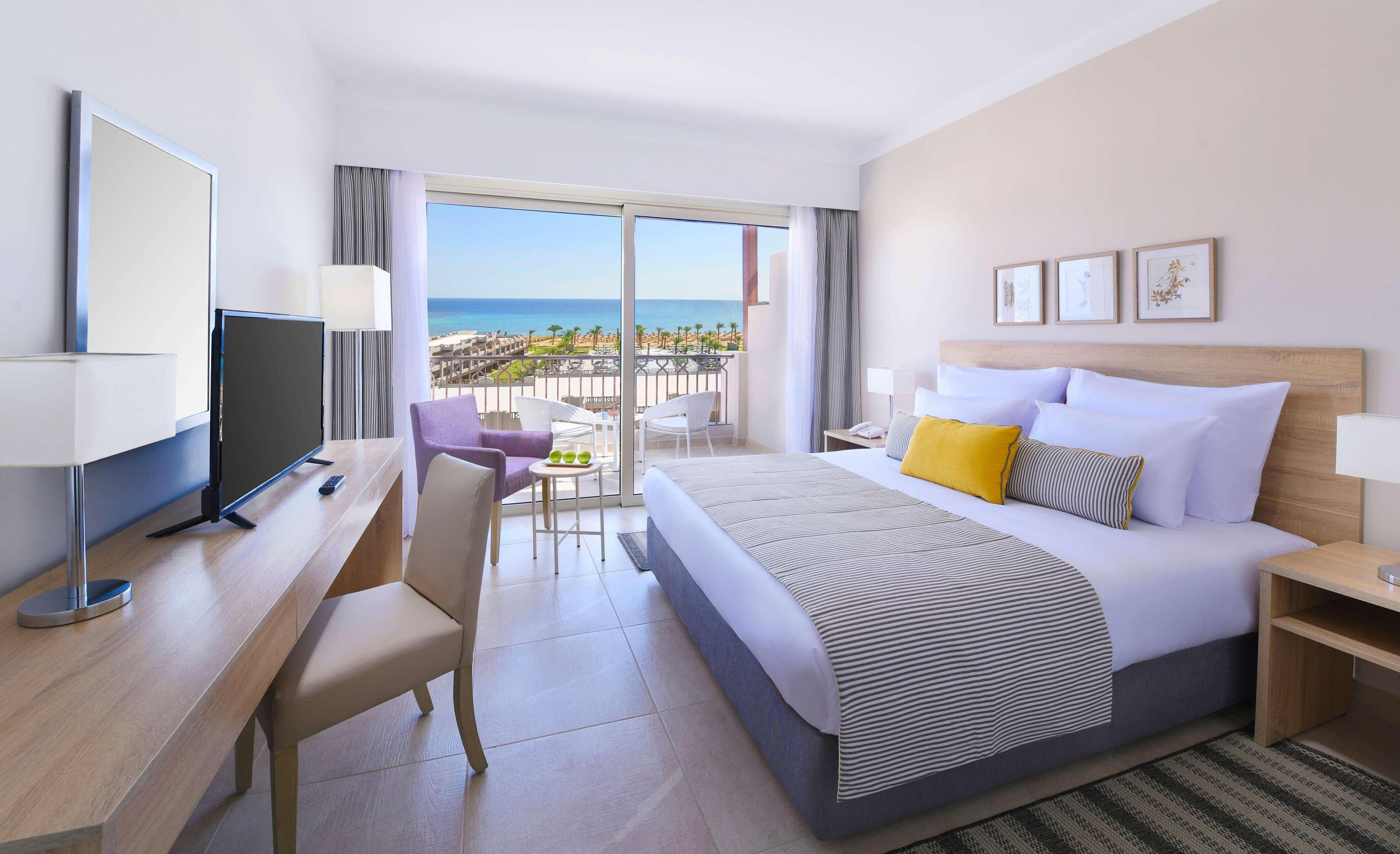Habitación de hotel con cama, escritorio, televisor, balcón con vista a la playa y ventana.