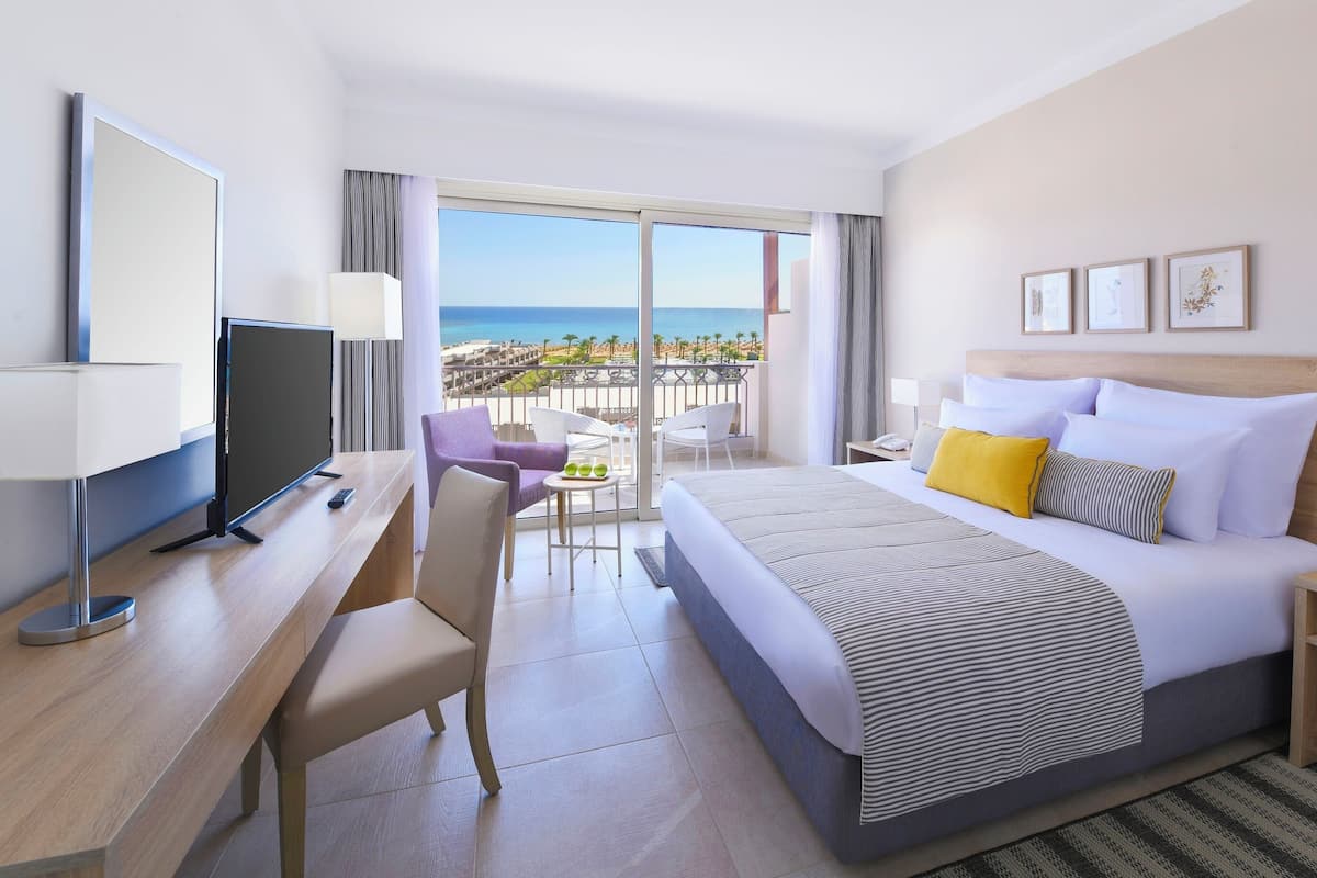 Ein Hotelzimmer mit Bett, Schreibtisch, Stuhl, Fernseher, einem Balkon mit Blick auf den Strand und einem Nachttisch mit Lampe.