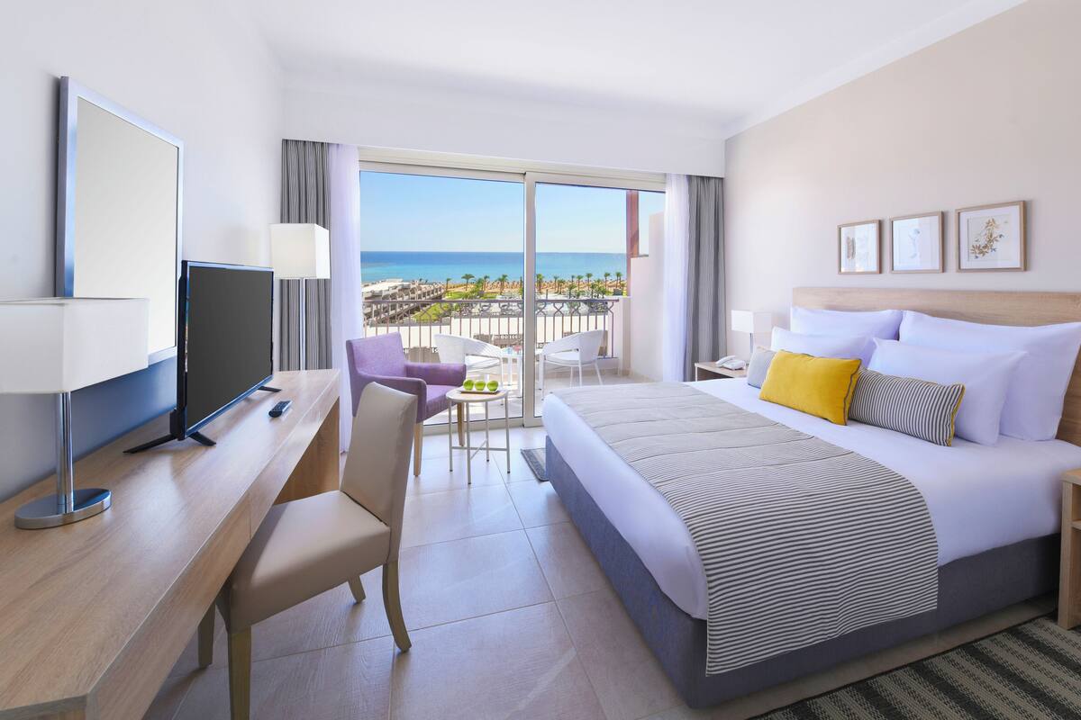 A hotel room with a bed, a desk, a TV, a balcony with a view of the beach.
