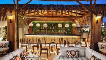 A cozy outdoor seating area with thatched roof, cushioned sofas, and a bar with a green wall display.