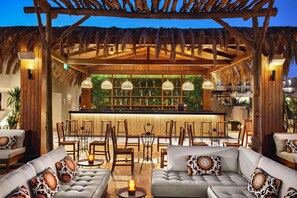 Een gezellig buitenverblijf met rieten dak, beklede banken en een bar met een groene wandpresentatie.