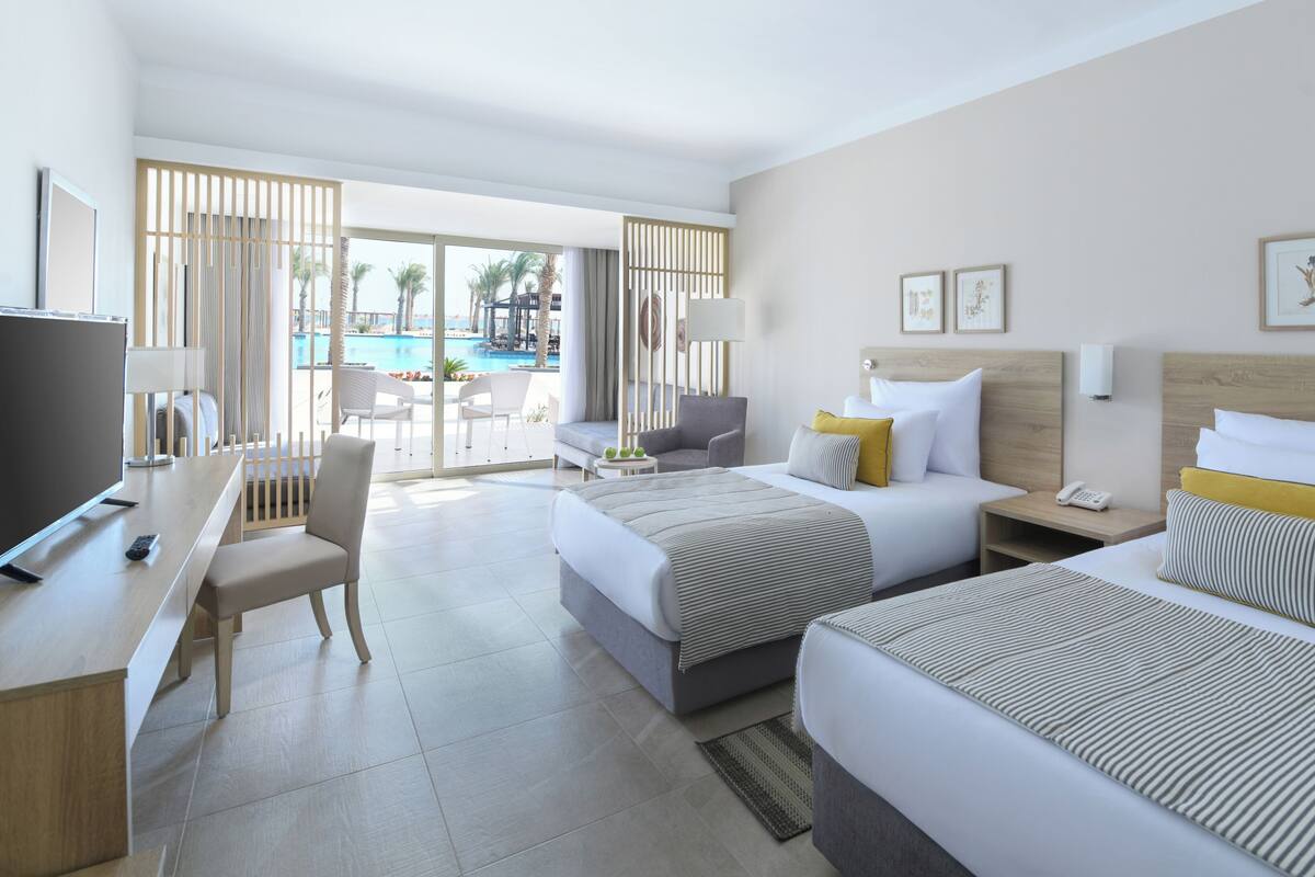 A hotel room with two beds, a desk, a chair, a TV, and a view of a pool.