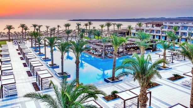 Un complexe hôtelier doté d’une piscine, de palmiers et offrant une vue sur la mer au coucher du soleil.