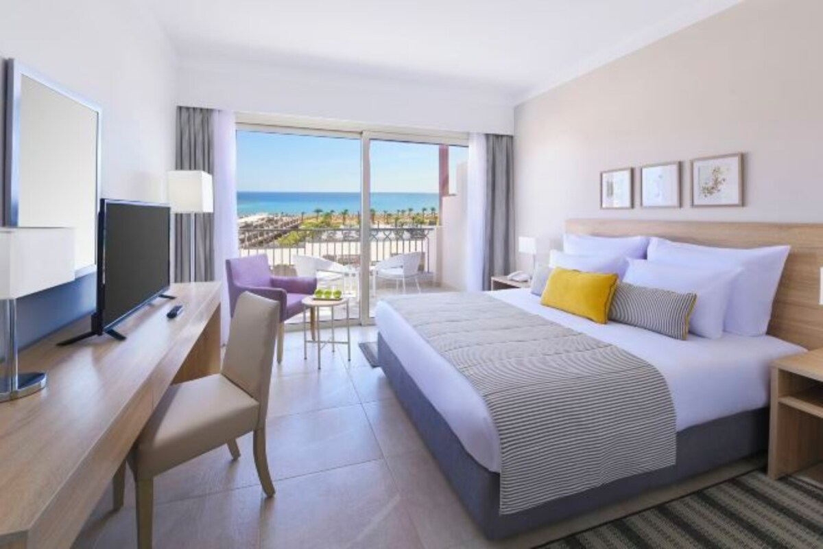 A hotel room with a bed, a desk, a television, a balcony with a view of the beach.