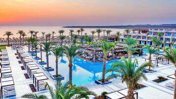 A resort with a pool, palm trees, and a view of the sea at sunset.