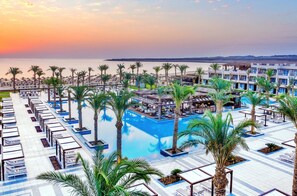 Een resort met een zwembad, palmbomen en uitzicht op zee bij zonsondergang.