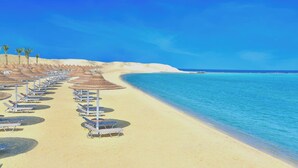 Une plage bordée de rangées de transats et de parasols, un ciel bleu azur et une mer calme.